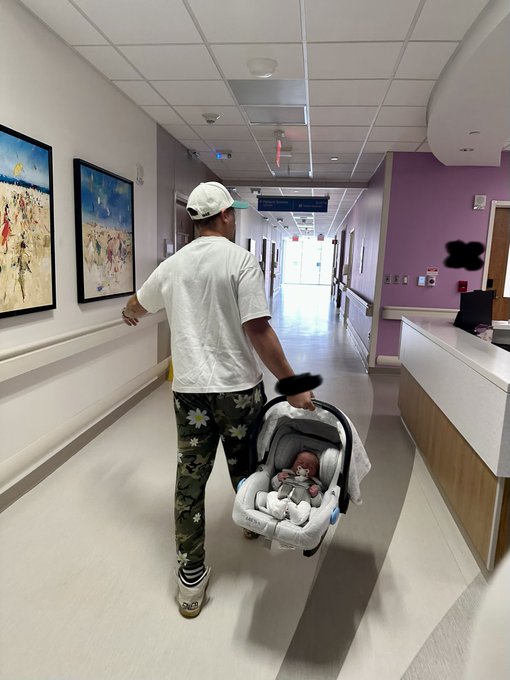 A man wearing a white t-shirt, cap, and floral pants walks down a hospital corridor while pushing a newborn baby in a car seat carrier.