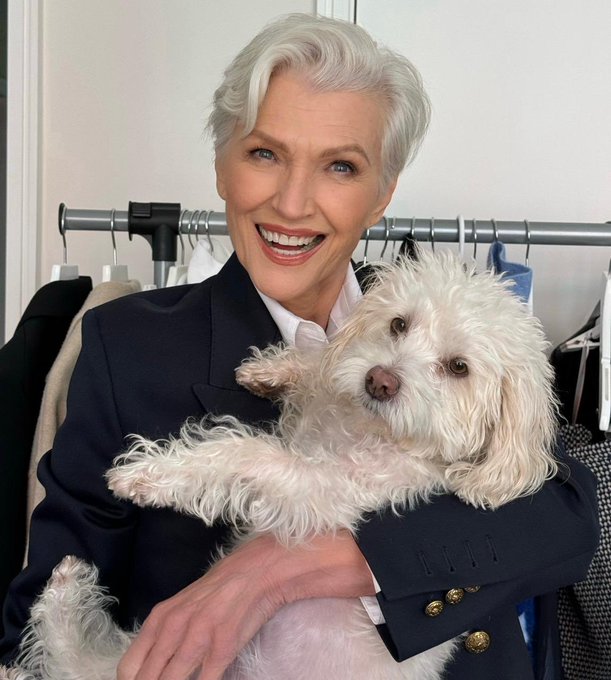 An older woman with short white hair smiles broadly while holding a fluffy white dog, standing in front of a clothing rack.
