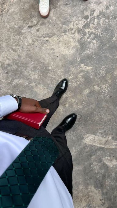 An overhead shot shows a person in black dress shoes and dark trousers holding a red notebook, with a green checkered folder nearby on a gray floor.