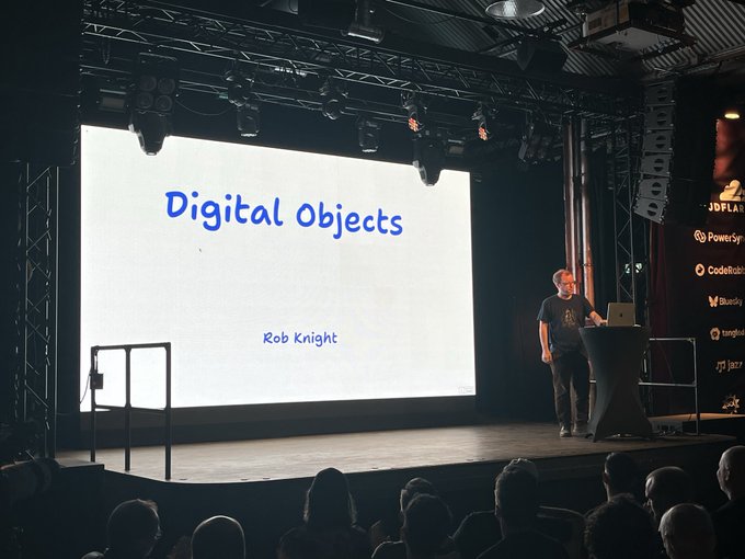A man giving a presentation on a stage next to a large screen displaying a slide that reads 'Digital Objects' by 'Rob Knight'.