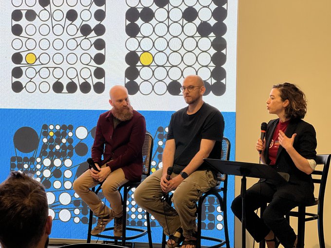 Three people sit on stools on a stage during a panel discussion, with the woman on the right speaking into a microphone against a colorful patterned backdrop.