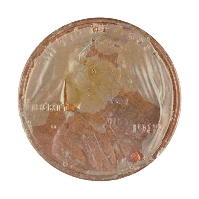 A heavily damaged and corroded 1938 Lincoln wheat penny, with a clear liquid or gel substance covering its surface, obscuring parts of the "IN GOD WE TRUST" and "LIBERTY" inscriptions.