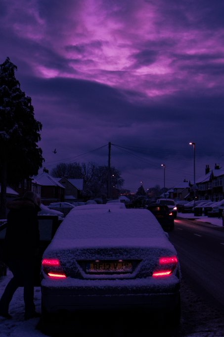 A car covered in snow parked on a street at night under a striking purple sky, with a person walking nearby.