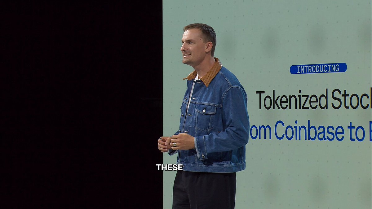 The image features a man standing next to a blue wall, wearing a denim jacket and smiling at the camera while giving a speech or presentation. The background of the image shows a white sign with black text that reads "→ 24/7 trading → Near-zero cost → Instant settlement → Available across the world Welcome to tokenized stocks on Base". This suggests that the man is discussing the concept of tokenized stocks, which are digital assets that can be bought and sold like traditional stocks. The image also contains a microphone, indicating that this could be an event or gathering where the speaker is addressing an audience.