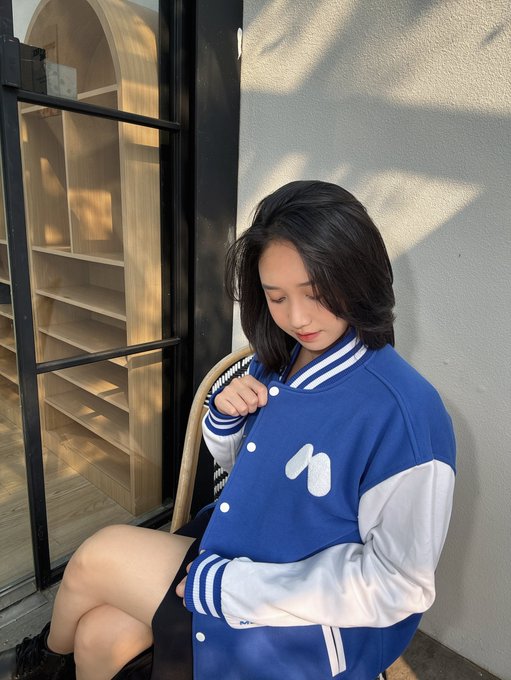 Young woman with short dark hair sitting by a window, wearing a blue and white varsity jacket with a heart logo and a checkered shoulder pattern.