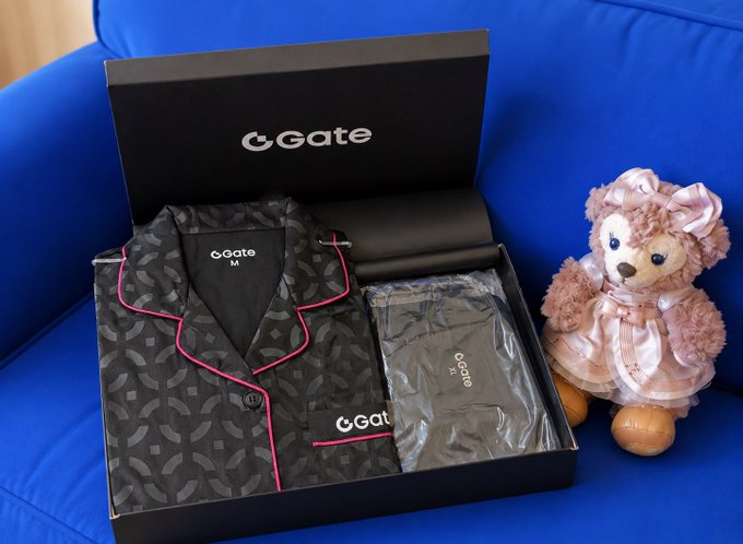 A photograph of a black Gate (CGate) branded gift box containing pajamas and a pouch, placed on a blue couch next to a pink teddy bear, likely merchandise from the Gate crypto exchange.
