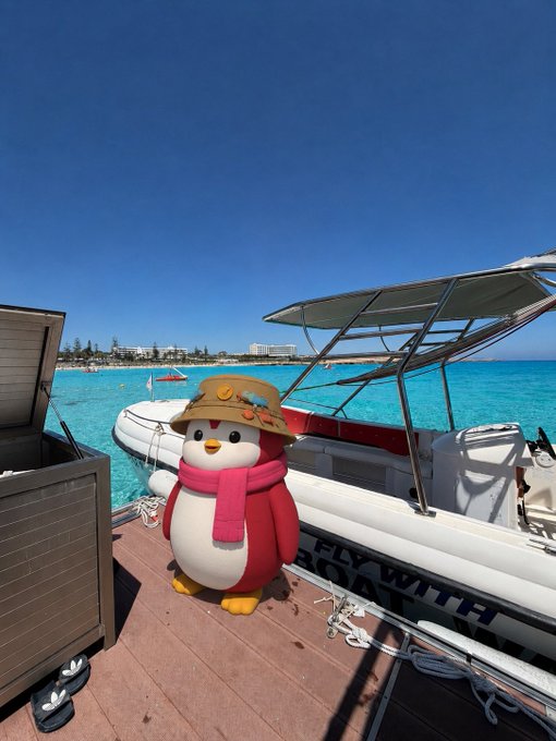 A cute penguin plush toy wearing a sun hat and pink scarf stands on a dock next to a boat with "FLY WITH" visible on its side, set against a turquoise ocean and clear blue sky. The serene tropical scene and forward-looking boat visually echo the optimistic post predicting Bitcoin's surge to $300,000 by 2026.