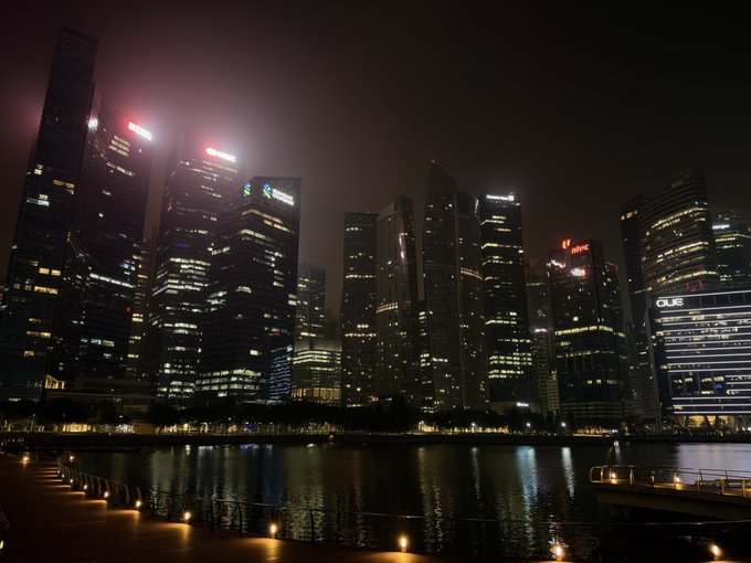A nighttime cityscape of a modern skyline featuring illuminated skyscrapers reflected in the water, with visible signage for OUE and other corporate buildings.