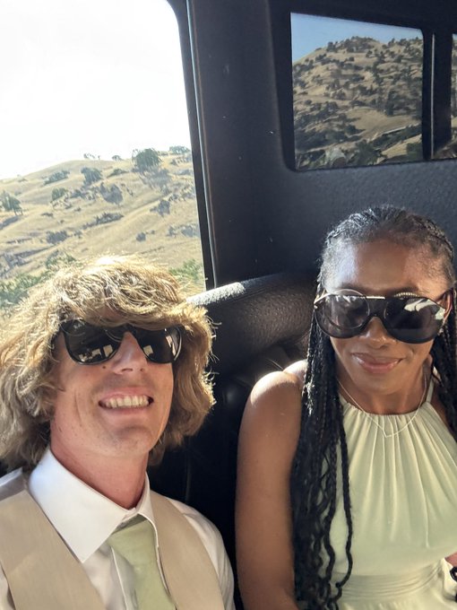 A smiling couple wearing sunglasses sits inside a vehicle with mountainous, dry terrain visible through the windows; the man wears a tan vest with a light green tie, while the woman wears a sleeveless cream top.