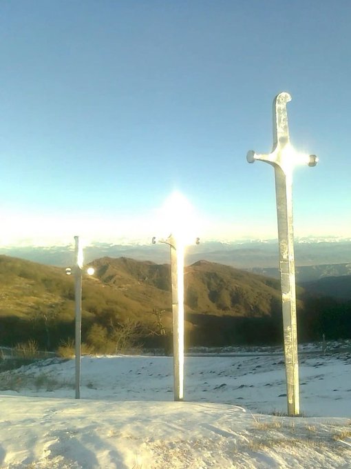 A scenic view of two large metal sword monuments planted in snowy ground, with mountains and a bright sun visible in the background.