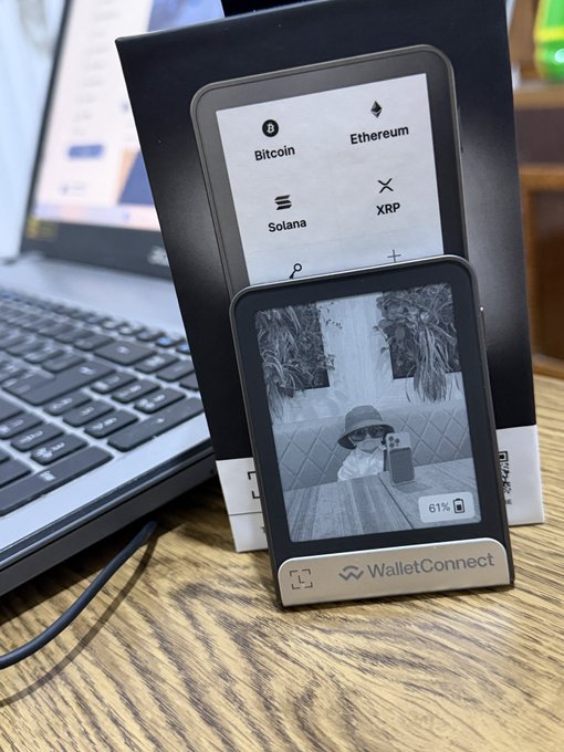 A device labeled 'WalletConnect' sits on a wooden desk next to a laptop, displaying a screen showing cryptocurrency options (Bitcoin, Ethereum, Solana, XRP) and a secondary screen showing a child at 61% battery.