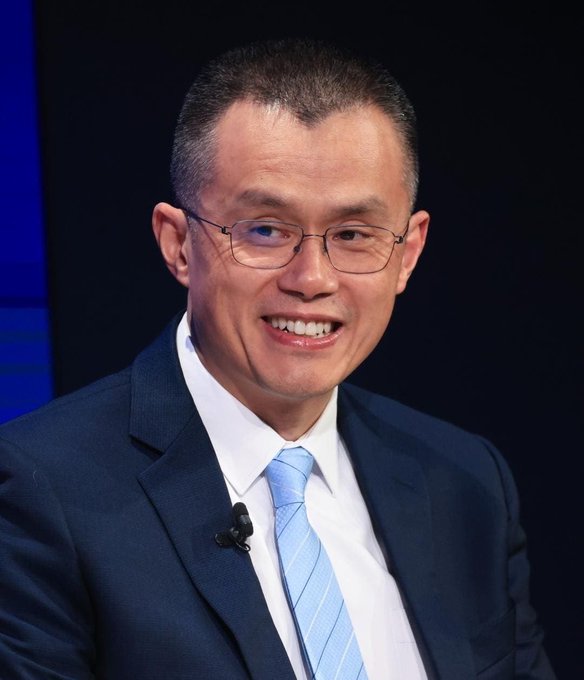 A smiling man wearing glasses, a dark suit, white shirt, and light blue tie with a lapel microphone appears to be speaking at an event, likely being CZ (Changpeng Zhao), the former CEO of Binance, in connection with the post's reference to choosing between 4,000 BNB tokens or spending a week with him.