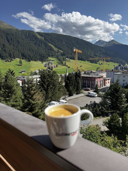 A coffee mug rests on a balcony railing overlooking an alpine village with construction cranes, lush green mountainsides, and a dramatic cloudy sky. The serene mountain view provides a calm backdrop that contrasts ironically with the post's request to share crypto wallet addresses—a classic setup often used in scam promotions designed to steal funds from unsuspecting followers.