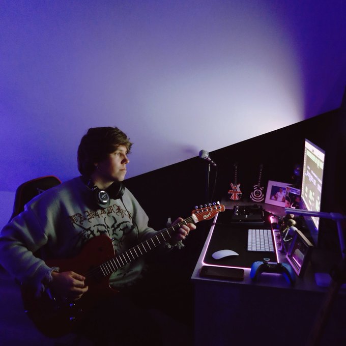 The image features a young man sitting at a desk with a guitar in front of him. He appears to be playing the guitar, possibly practicing or enjoying his hobby. The room has a purple wall behind him, which adds a unique color to the scene. There is also a computer monitor and keyboard on the desk, indicating that he might use them for music production or other creative purposes.