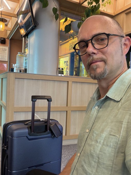 A selfie of a bald man with glasses and a goatee wearing a green button-up shirt, standing next to a dark blue rolling suitcase in what appears to be a hotel lobby or cafe.