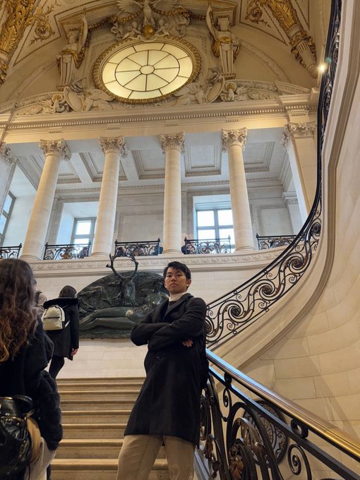 A person with crossed arms stands on an ornate, grand marble staircase with classical columns and decorative sculptures in the background.