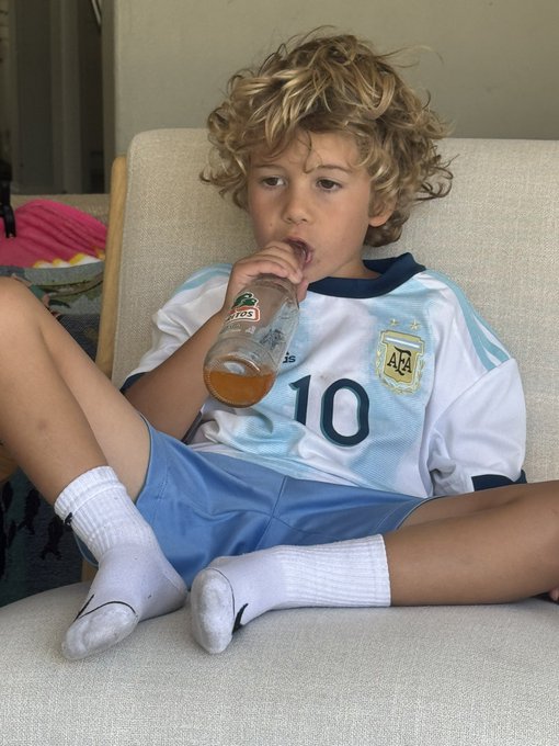 A curly-haired boy wearing an Argentina national team soccer jersey with the number "10" and AFA crest sits on a couch sipping Jarritos soda, casually chilling just like the laid-back "chimp" vibe of the post.
