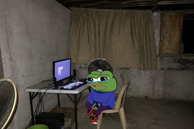 A Pepe the Frog cartoon character wearing a blue hoodie and sneakers sits on a white plastic chair at a desk with a laptop displaying a purple screen, next to an electric fan in a concrete-walled basement.