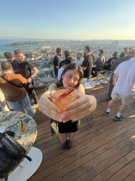 The image shows a woman holding an orange drink, possibly a cocktail or a fruity beverage, with her arms outstretched as if she's posing for a picture. The setting appears to be at a rooftop party or event, where people are enjoying themselves in the background. There is also a handbag visible on the ground near one of the guests.