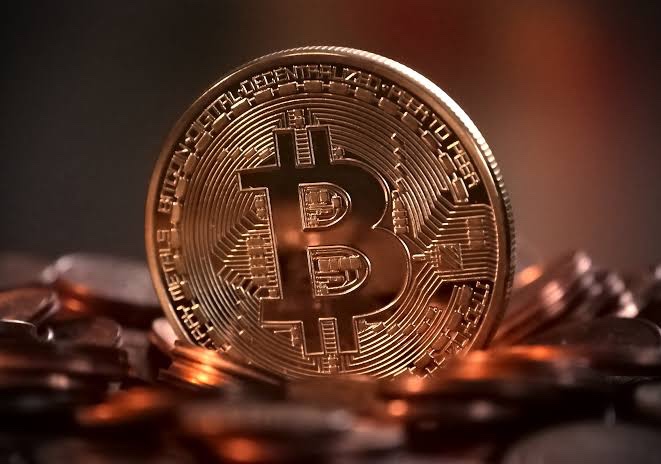 A shiny gold Bitcoin coin stands upright atop a pile of other coins, displaying the iconic "₿" symbol and the inscription "BITCOIN DIGITAL DECENTRALIZED PEER TO PEER" along its edge, symbolizing Capital B's plan to expand its Bitcoin treasury holdings.