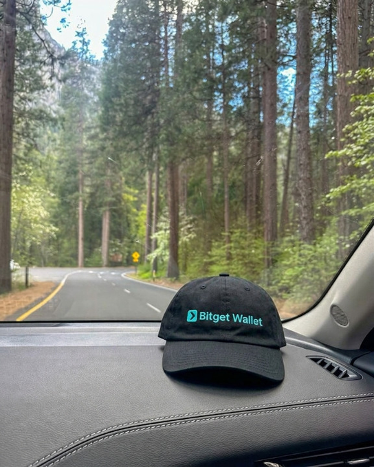 A black "Bitget Wallet" cap rests on a car dashboard, framed against a sunlit forest road ahead—capturing the "fresh vibes, secure wallet" mood of the post as the journey begins.