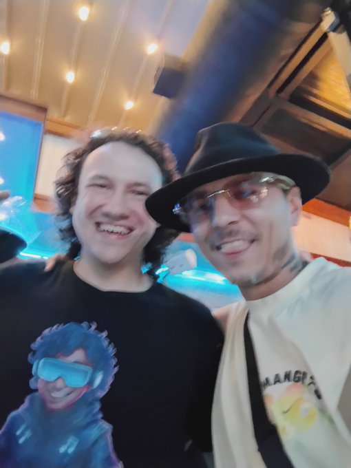 Two men smile for a close-up selfie at a lively event with blue lighting, one wearing a black t-shirt with a cartoon graphic and the other in a black hat, sunglasses, and a white 'HANG' t-shirt.