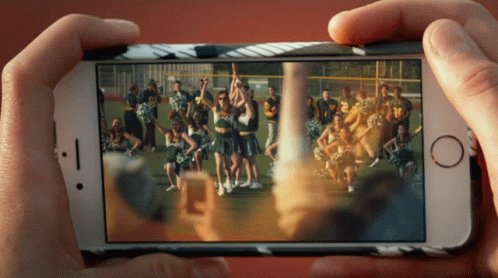 The image shows a person holding up their phone, displaying an image of a group of cheerleaders performing on a field. The viewer's perspective is from behind the phone screen, looking at the cheerleaders as they dance. The focus of the image seems to be on capturing and sharing this moment with others through social media or other platforms.