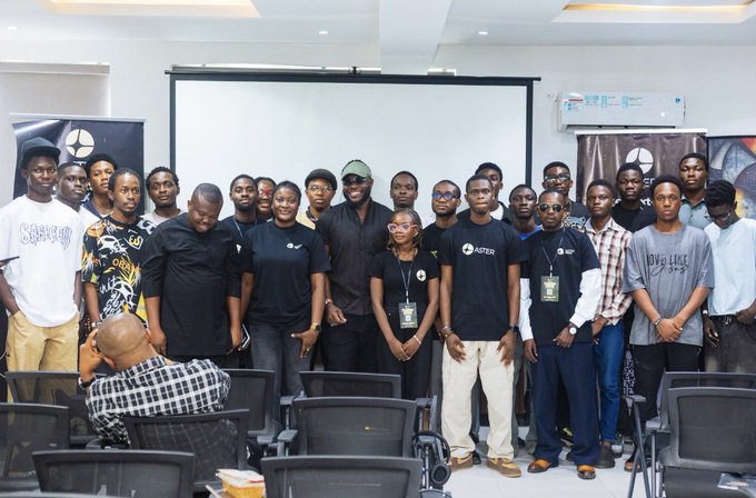 A group photo of approximately twenty people, mostly young men in black shirts, posing together indoors in what appears to be a classroom or workshop setting.