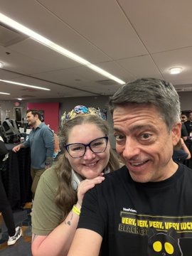A selfie of a smiling man in a black 'Werk Werk Werk Werk Blacklist' t-shirt posing with a woman wearing glasses and a colorful headband at an indoor event.