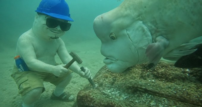An underwater scene showing a small figurine of a man wearing a blue hard hat and sunglasses, holding a hammer, facing a large fish in murky green water.