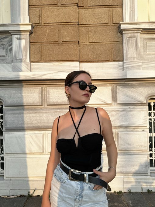 A young woman wearing a black strappy top, light-wash jeans, sunglasses, and a choker poses against a classical stone building in warm sunlight. The "Gm friends" caption suggests a cheerful, stylish weekend greeting showcasing her trendy outfit.