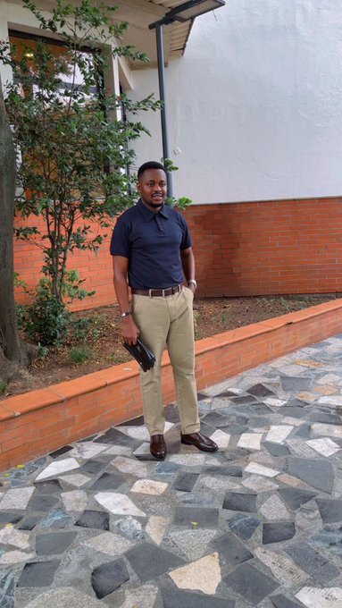 A young man wearing a navy polo shirt and beige trousers stands on a decorative stone pathway in front of a building with brick walls and greenery.