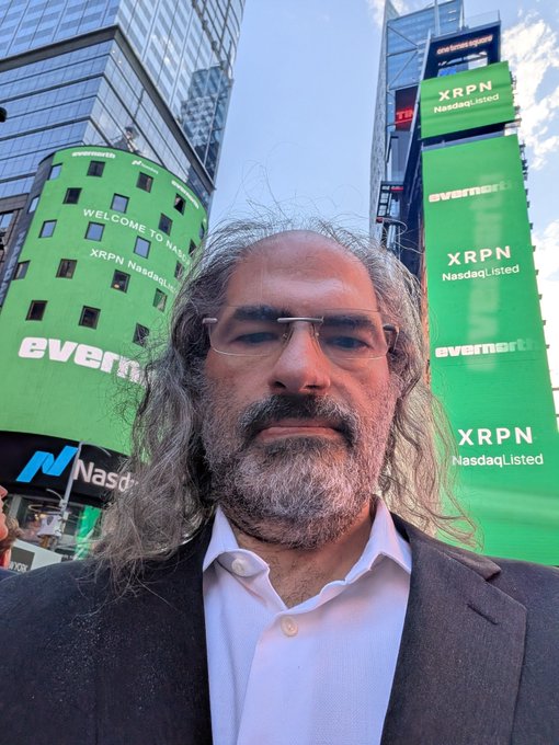 A man with long gray hair and glasses stands in Times Square in front of bright green NASDAQ billboards displaying "XRPN NasdaqListed" and "evermarkit," with one screen reading "WELCOME TO NASDAQ XRPN NasdaqListed," celebrating XRP's listing on the Nasdaq—visually echoing the post's bullish "big green candle" prediction.
