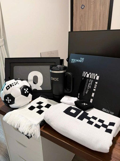 A collection of OKX VIP merchandise including a soccer ball, towel, scarf, tumbler, blanket, and a book titled "The New Money App" displayed on a wooden surface.
