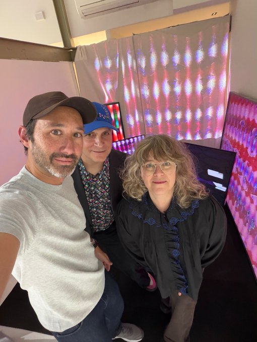Three people pose for a selfie at what appears to be an art exhibit with illuminated pink and purple panels in the background. The man on the left wears a brown cap and gray shirt, while the woman on the right wears glasses and a black ruffled blouse.
