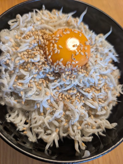 A black bowl filled with small white fish (likely whitebait or similar) topped with sesame seeds and a raw egg yolk in the center.