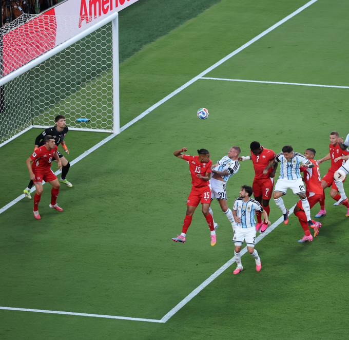 A soccer match scene with players in red and white/light blue striped jerseys, with the ball in the air near the goal.