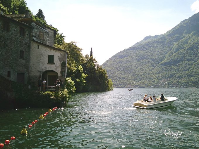 The image shows a group of people enjoying themselves on a boat near a mountain range. The boat appears to be a motorboat with several passengers onboard. In the background, there are two buildings located close to the water's edge, possibly houses or small businesses. A person is standing next to one of these buildings, and another individual can be seen further away from the scene.