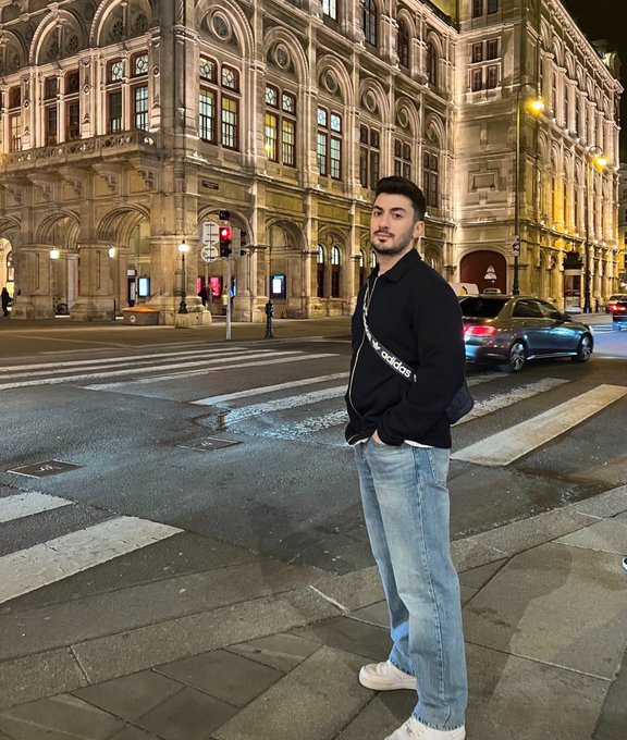 A stylish young man stands on a city crosswalk at night, wearing a black Adidas track jacket and light blue jeans, with an illuminated historic building and a parked car behind him—setting the scene for a cheerful "Good morning" greeting despite the evening setting.