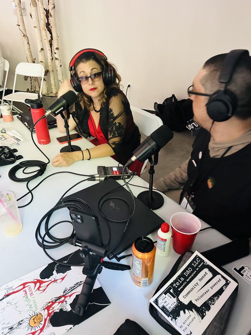 Two people wearing headphones sit at a table with microphones, recording a podcast episode for 'Helladuo LLC.'