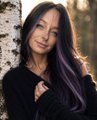A portrait of a woman with long, dark hair featuring purple highlights, wearing a black long-sleeved top, smiling gently while leaning against a tree.