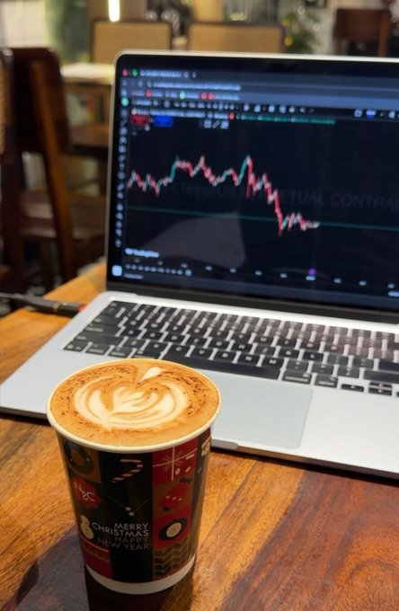A latte with intricate foam art sits beside an open laptop displaying a stock chart labeled "BITCOIN ANNUAL FUTURES CONTRACT," capturing a moment of remote work from a café. The "Merry Christmas Happy New Year" cup and portable trading setup perfectly embody the freedom of working anywhere.