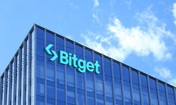 A modern glass office building displaying the 'Bitget' logo in teal against a blue sky.