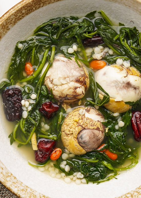 A bowl of soup contains green leafy vegetables, whole eggs, red dates, goji berries, and small white grains in a clear broth.