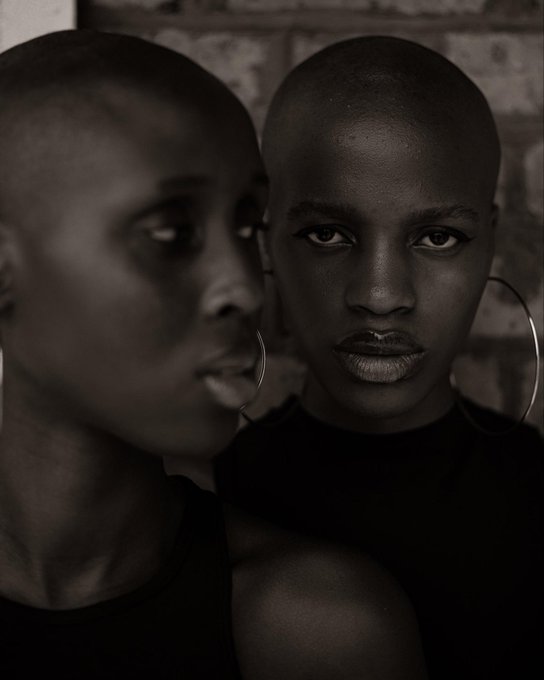 A black and white portrait of a woman with a shaved head and large hoop earrings, appearing twice in the frame against a brick wall background.