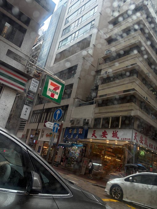 A rainy street view through a wet car windshield showing a 7-Eleven convenience store sign and Chinese storefronts in a dense urban area, likely Hong Kong.