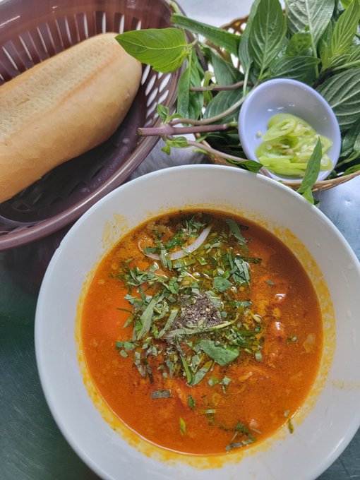 A bowl of red curry soup garnished with fresh herbs, accompanied by a basket of crusty baguette, fresh mint and basil leaves, and a small dish of sliced green chilies.