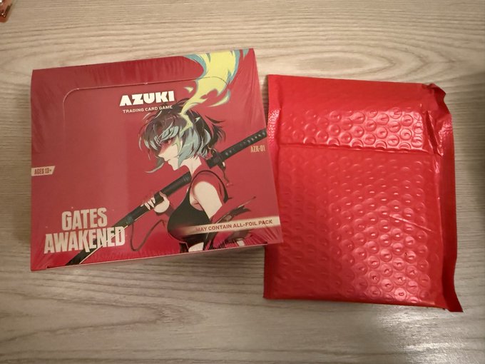 An unopened Azuki TCG "Gates Awakened" booster box (ALX-01, ages 13+) sits next to a red bubble mailer on a wooden surface, ready to be opened and ripped. The colorful anime-style box art features a character with teal hair holding a sword, matching the excited anticipation of the post.