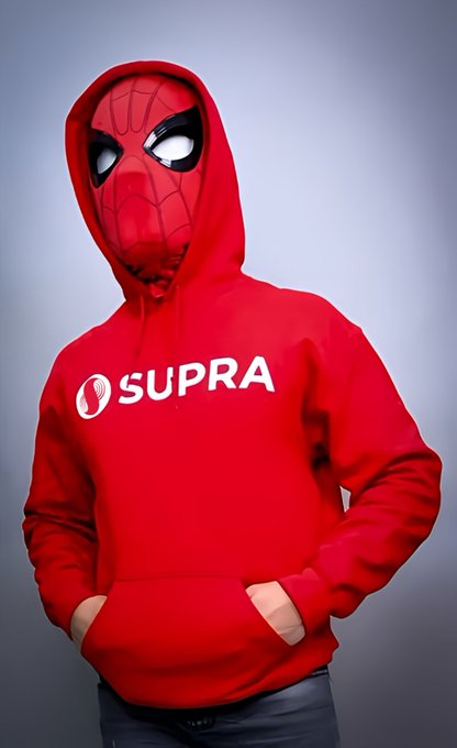 The image features a person wearing a red hoodie that says "SURRA" on it. The individual appears to be posing for a photo, with their hands in their pockets against a gray background. The caption of the image reads "GM CT It' s a great day to be obsessed with $SUPRA. Explore the ecosystem, support the creators, connect with the community."