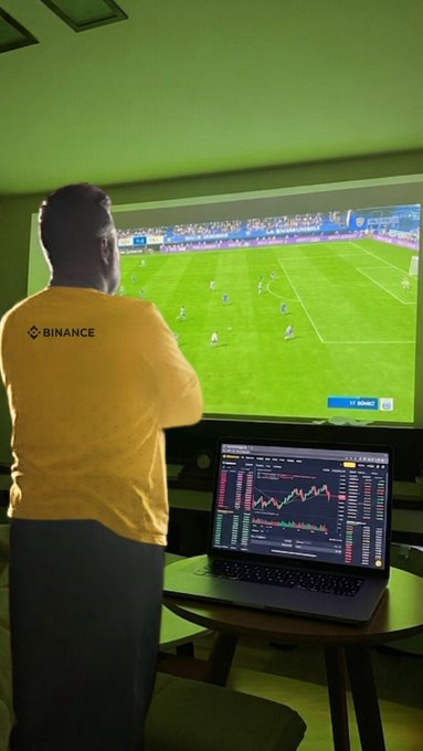 A person wearing a yellow "BINANCE" t-shirt watches a World Cup soccer match on a large TV while simultaneously monitoring cryptocurrency trading charts on a laptop. The setup humorously illustrates how traders multitask by keeping an eye on both sports games and live market activity at the same time.